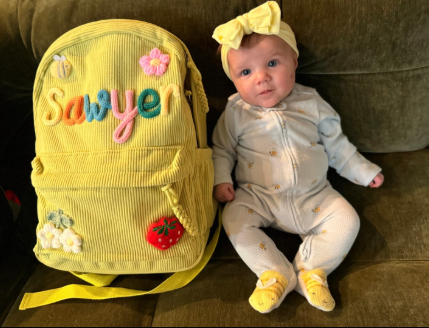 Baby Sawyer next to a yellow corduroy backpack with her name embroidered
